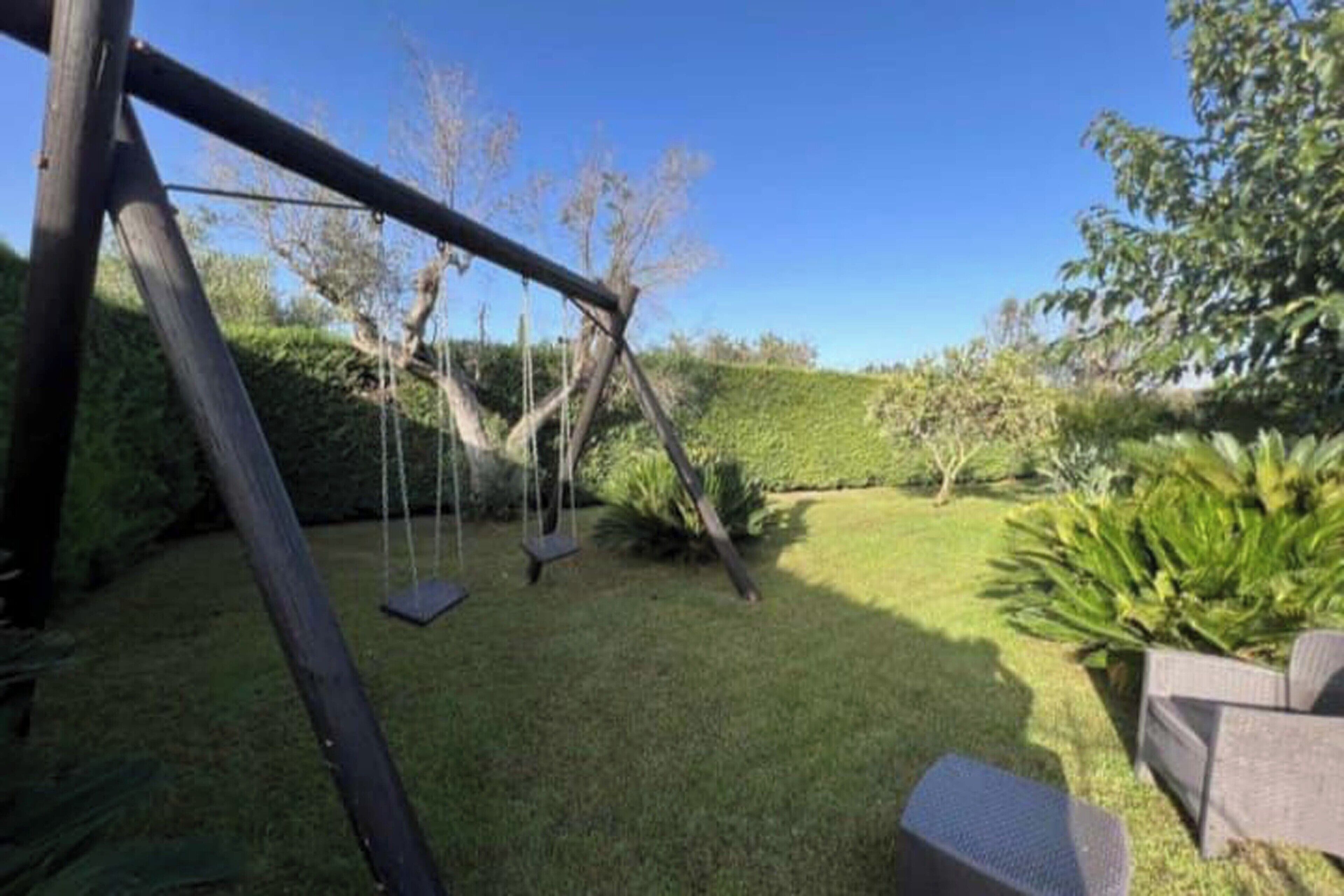 Ein hölzernes Schaukelgestell in einem gepflegten Rasenstück, mit einer Hecke und einem Stuhl.