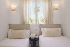 A room with two beds, each with a headboard and a pillow, a small table with two vases, and a window with curtains. - Villa Rosa primera linea (Conil de la frontera)