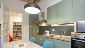 Eine kleine Küche mit einem Holztisch, blaugrünen Stühlen, einem Einbauofen und einem Bücherregal im Hintergrund.