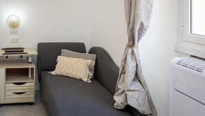 Ein kleiner, gemütlicher Raum mit einer grauen Couch, einer gemusterten Decke, einer Wandlampe und einem Fenster mit Vorhang.