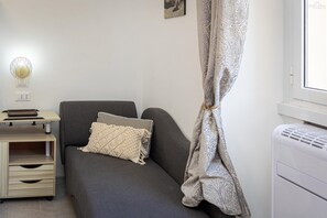 Ein kleiner, gemütlicher Raum mit einer grauen Couch, einer gemusterten Decke, einer Wandlampe und einem Fenster mit Vorhang.