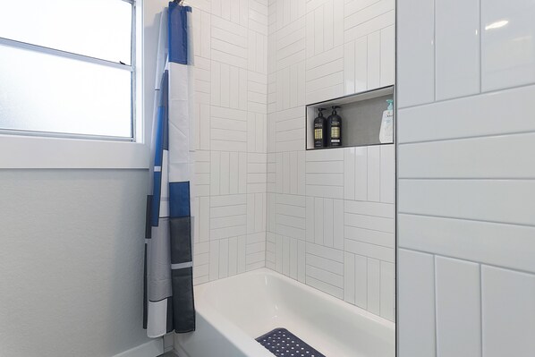 Un baño con paredes de azulejos blancos, una bañera, un cortinón de ducha y un estante con artículos de aseo.