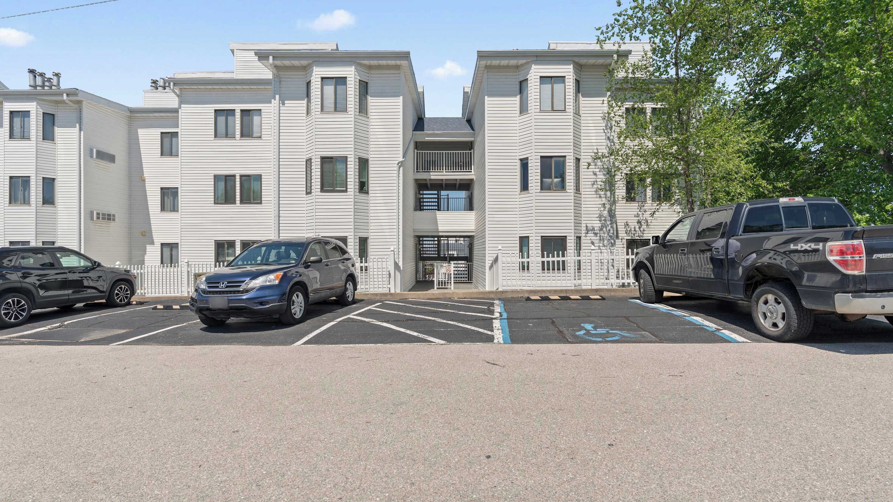 Un parking avec trois véhicules, un bâtiment doté d’une entrée centrale et une clôture blanche.