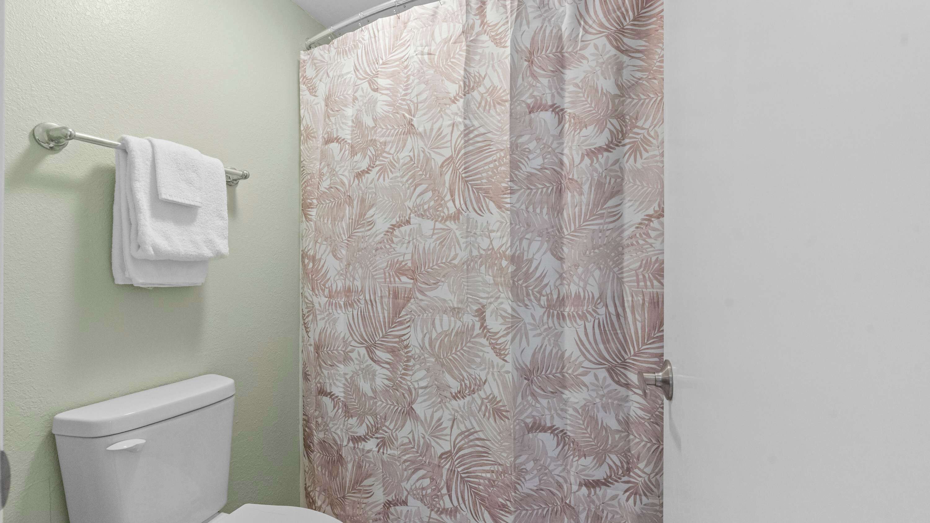 Een badkamer met een toilet, een handdoekrek met een opgevouwen handdoek, en een douchegordijn met een bladmotief.