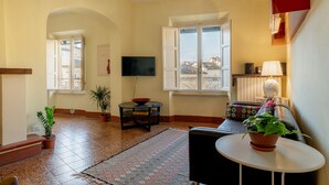 Een woonkamer met een televisie, bank, tafel, potplanten en uitzicht door een raam.