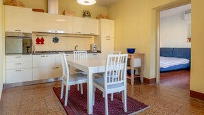 Een kleine keuken met witte kasten, een eettafel met stoelen, een kleine koelkast, en een blauwe kom op een bijzettafeltje.