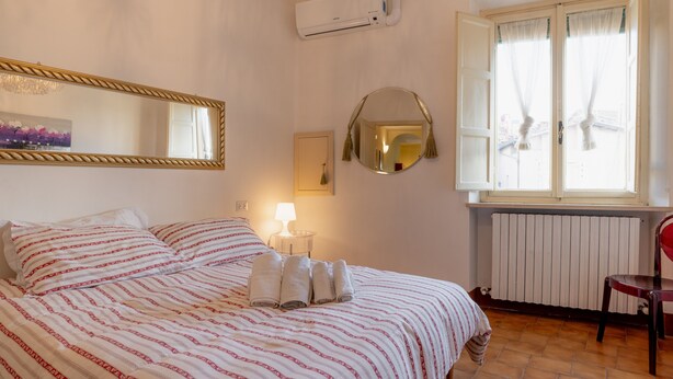 Een net opgemaakt bed met gestreept beddengoed, een ronde spiegel, een wandairconditioner, een raam met gordijnen, en een radiator.