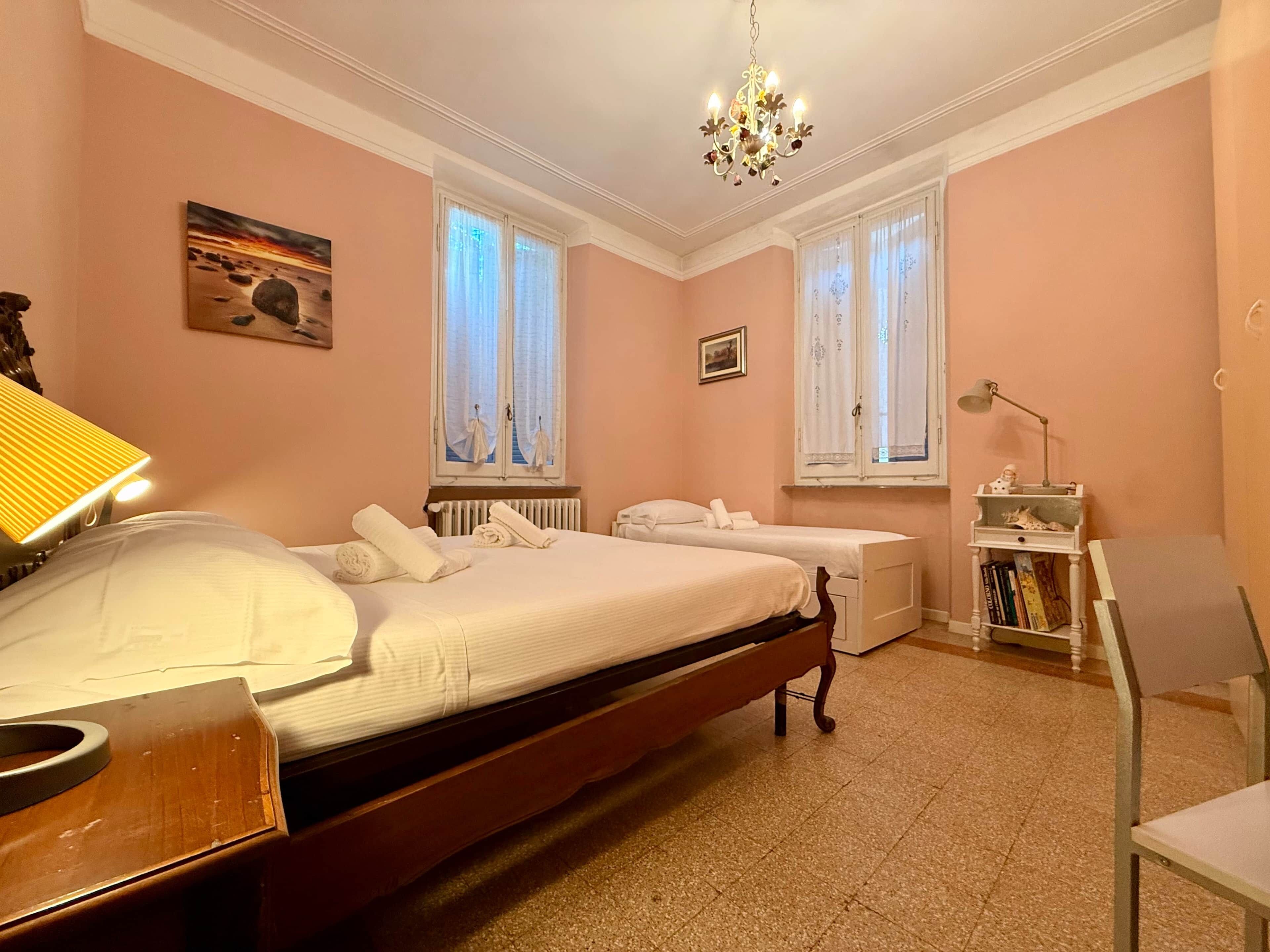 Ein Schlafzimmer mit einem großen Bett, zwei mit Vorhängen versehenen Fenstern, einem Heizkörper, einem Nachttisch, einer Lampe, einem Bücherregal und einer Deckenleuchte.