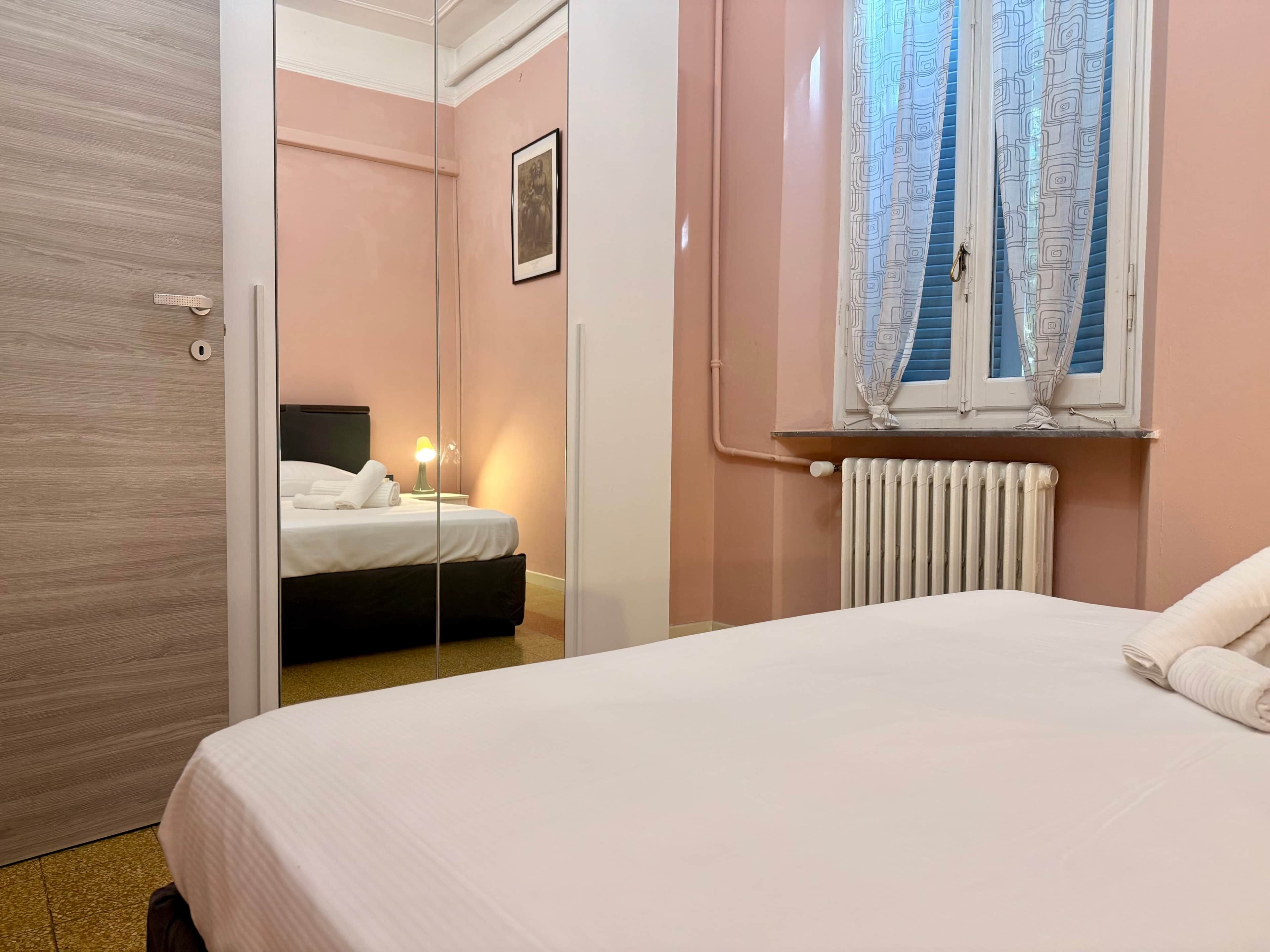 A bedroom with a bed, a bedside table with a lamp, a radiator, and a window with patterned curtains.