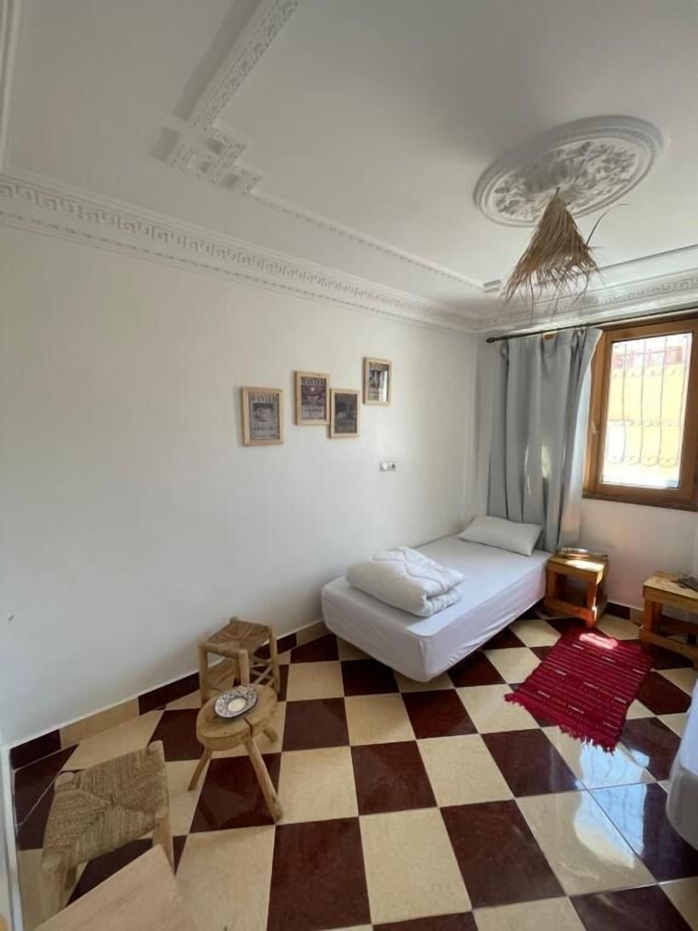 Una habitación con piso a cuadros, una cama con colchón y almohadas blancas, un taburete de madera, un tapete rojo y un ventilador de techo.