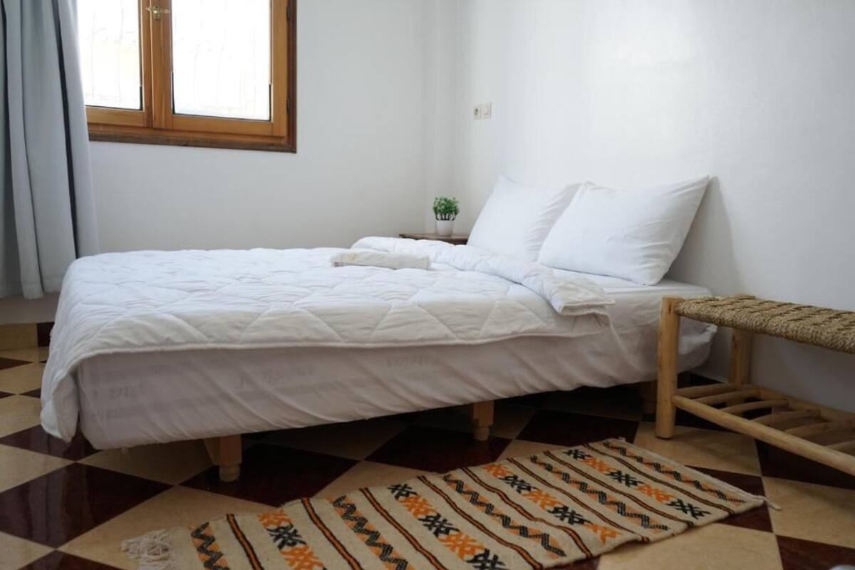 Ein ordentlich bezogenes Bett mit weißer Bettwäsche, ein kleiner Nachttisch mit einer Pflanze und ein karierter Boden.