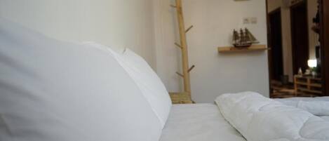 Una cama bien hecha con ropa blanca, un perchero de madera y un pequeño estante con un modelo de barco.