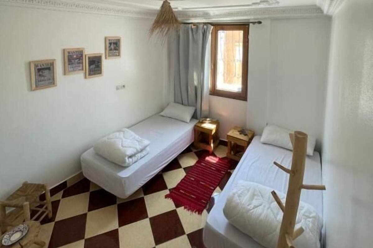 Ein kleiner, hell erleuchteter Raum mit zwei Einzelbetten, einem Schachbrettboden, einem Fenster mit Vorhängen und einer Decke mit dekorativer Stuckleiste.