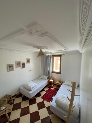 A small, well-lit room with two single beds, a checkered floor, a window with curtains, and a ceiling with decorative molding. - Epicswellhouse (Aourir)