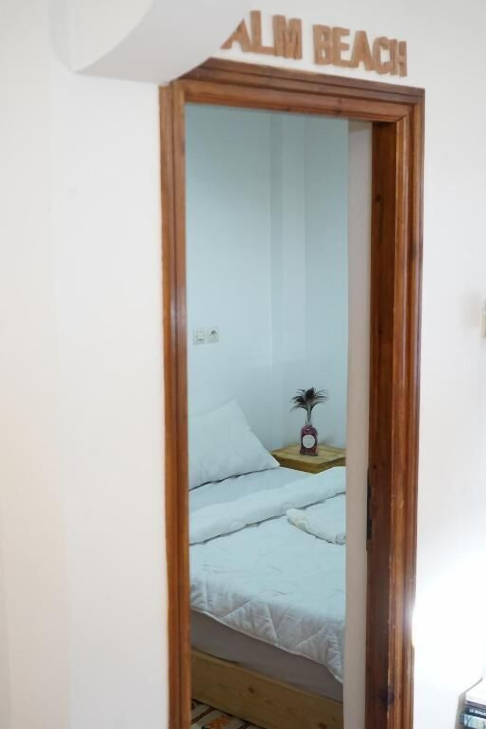 Une chambre avec un lit, une petite table de chevet sur laquelle se trouve un vase, et une pancarte au-dessus de la porte indiquant « ALM BEACH ».