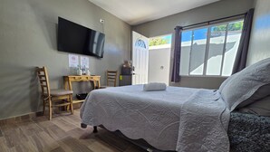 A hotel room with a bed, a desk, a chair, a TV mounted on the wall, and a window with curtains.
