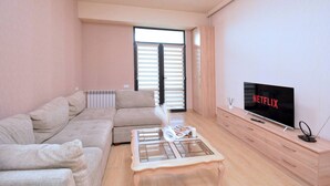 En moderne stue med en beige sofa, et sofabord med glasplade, et tv-bord i træ og et stort vindue med persienner.