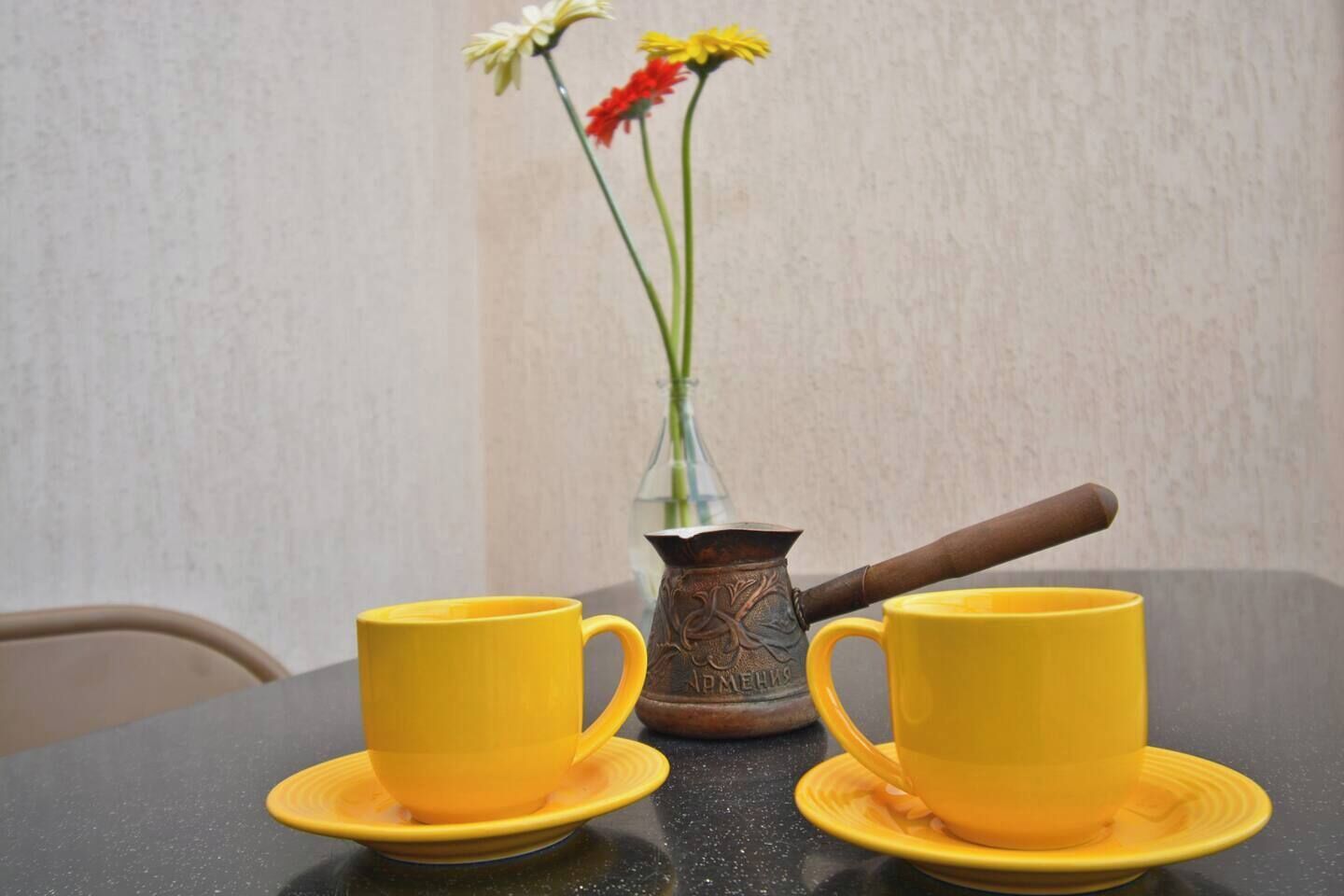 Twee gele kopjes op schoteltjes, een metalen karaf en een vaas met bloemen op een tafel.