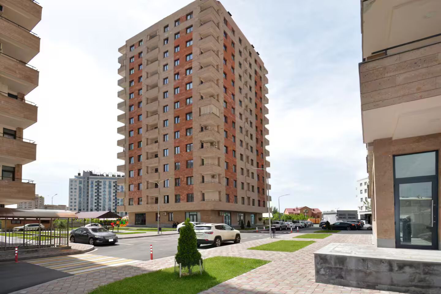 A high-rise residential building with multiple balconies, a paved pedestrian area, and parked cars.