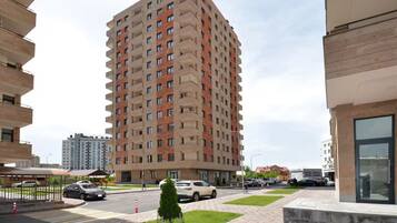 A high-rise residential building with multiple balconies, a paved pedestrian area, and parked cars.