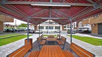 A covered outdoor seating area with benches and tables, surrounded by a paved walkway and a parking lot.