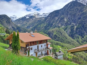 Ein zweistöckiges Holzhaus mit Balkon, umgeben von üppigem Grün und Bergen im Hintergrund.