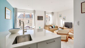Eine moderne Küche mit einer großen Kochinsel, ein Essbereich mit einer Couch und Fernseher sowie große Fenster mit Blick auf die Stadt.