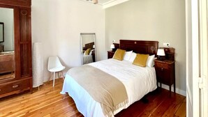 Une chambre avec un plancher en bois, un lit avec une literie blanche et beige, une table de chevet avec une lampe, un miroir et une armoire en bois.