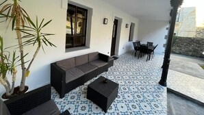 Un espace extérieur abrité comprenant un canapé, des fauteuils et une table. On y trouve également des plantes en pot et un sol carrelé.