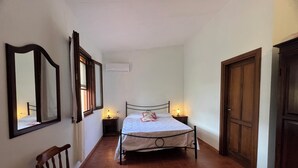 Un couloir étroit avec un lit, deux tables de chevet avec des lampes, une chaise en bois et un grand miroir sur le côté gauche.