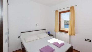 Ein Schlafzimmer mit Bett, einem Fenster mit Vorhängen, einem Nachttisch und Blick aufs Meer.