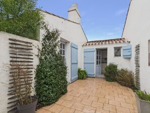 Une cour avec un chemin pavé, un bâtiment blanc aux volets bleus et une plante en pot.