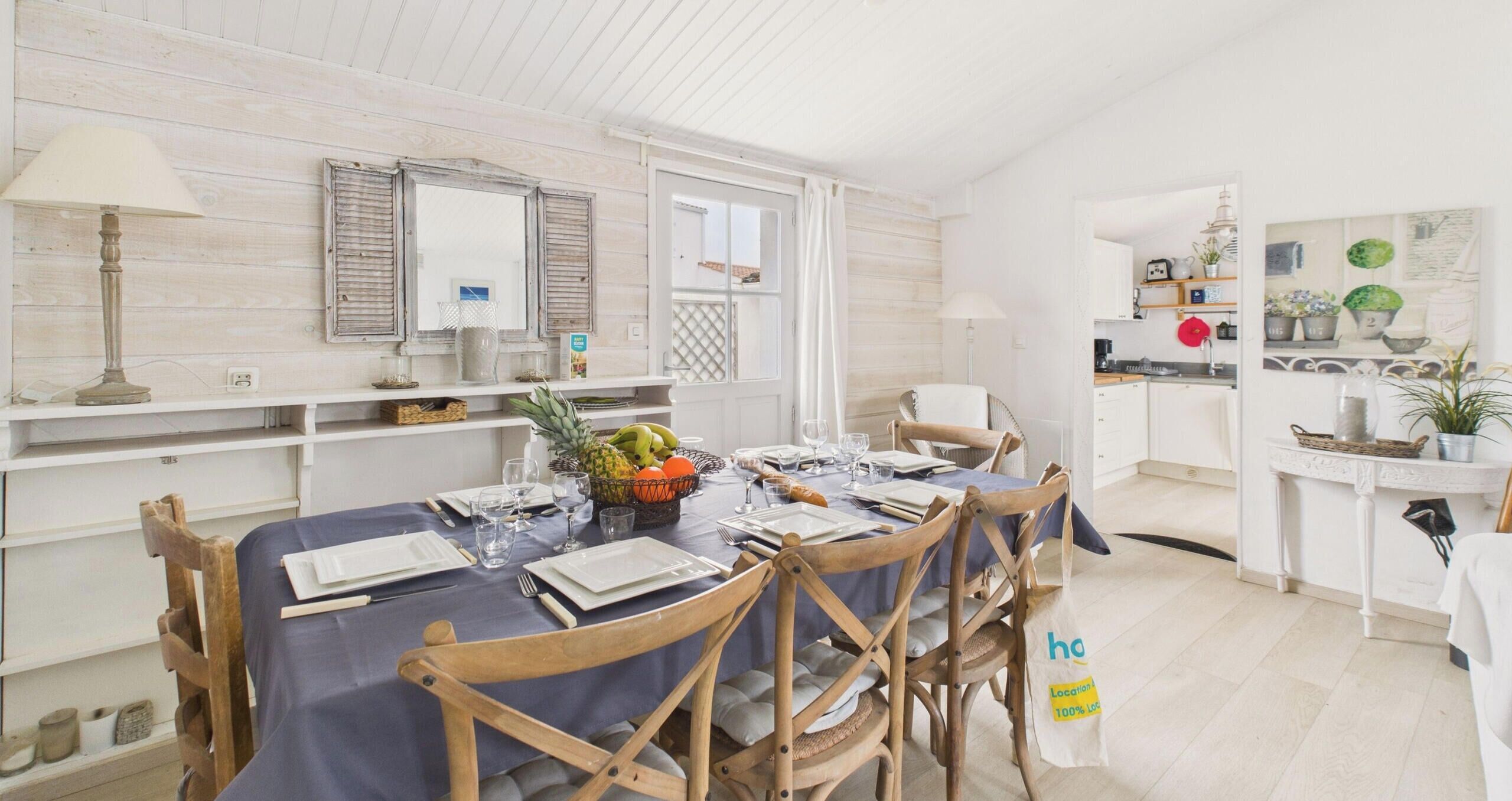 A dining area with a table set for four, wooden chairs, a fruit basket, and a white cabinet with kitchenware.
