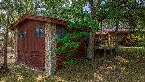 Une petite cabane avec des fondations en pierre et des murs en bois, entourée d’arbres et d’une zone herbeuse.