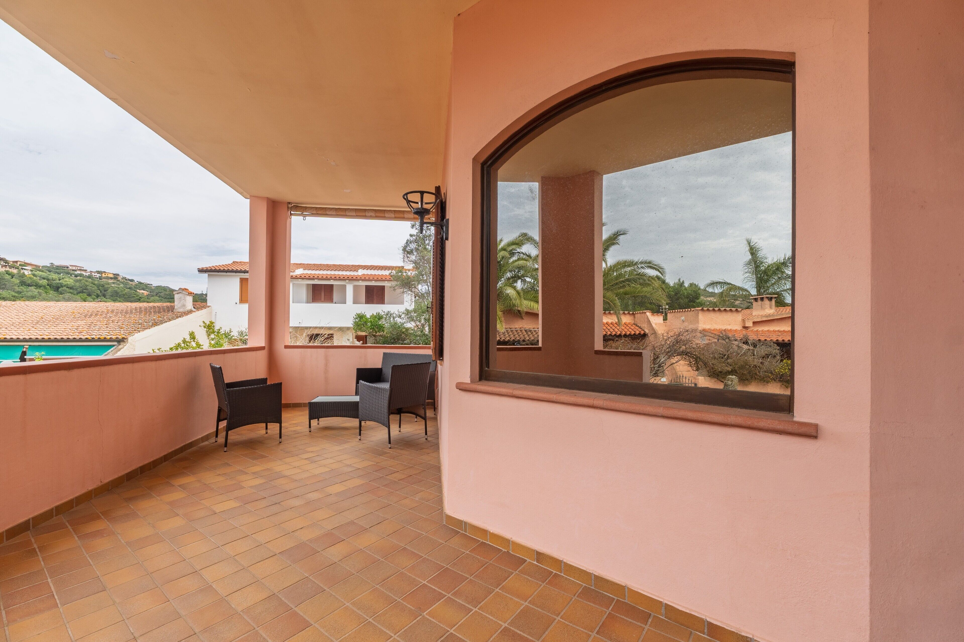 A balcony with tiled flooring, a chair, a table, and a window overlooking a pool and greenery.