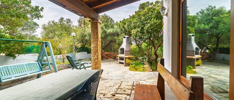 An outdoor patio with stone flooring, wooden furniture, and a view of a garden and trees.