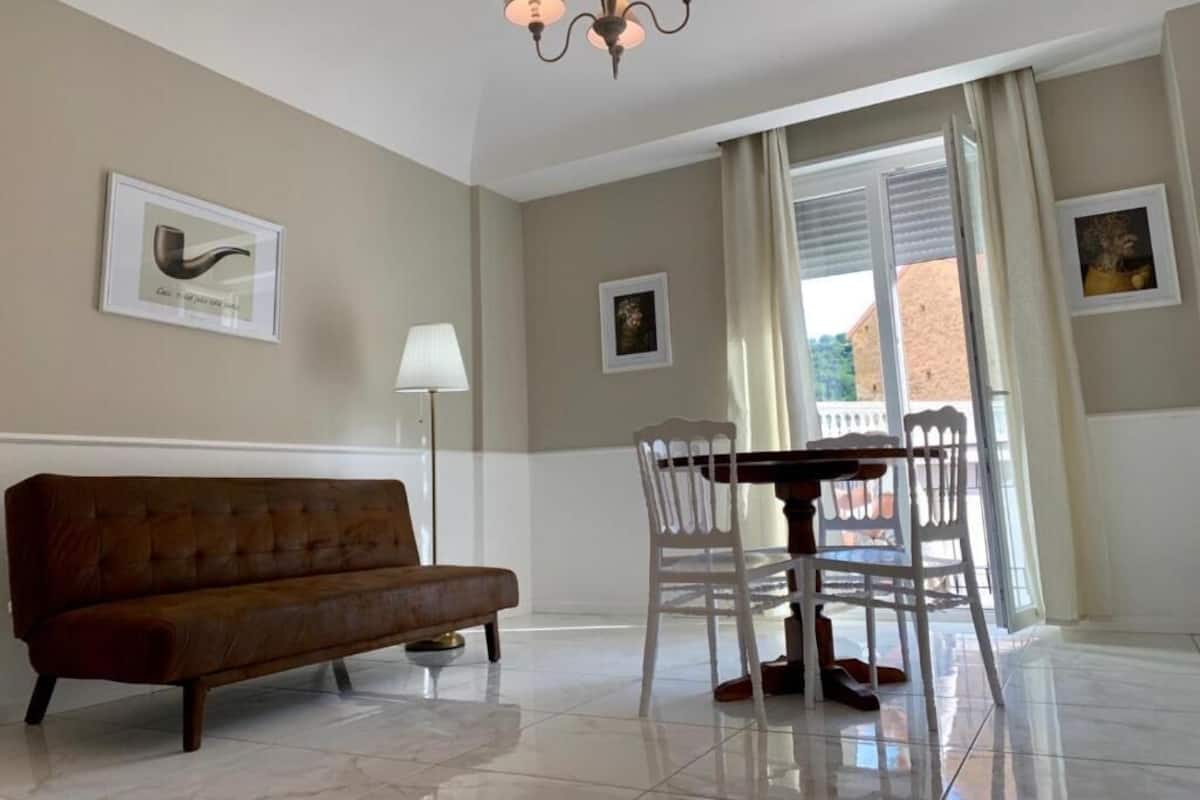 Ein Zimmer mit einem braunen Sofa, einem Esstisch mit Stühlen, einer Chandelüster und gerahmten Bildern an den Wänden.