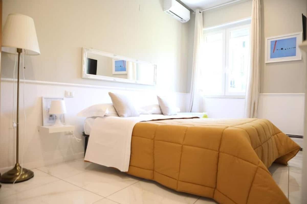Ein Hotelzimmer mit Bett, Nachttisch mit Lampe, Fenster mit Vorhängen und einer Wand-Klimaanlage.
