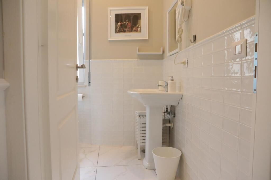 Un baño estrecho con azulejos blancos, un lavamanos pedestal y un pequeño cubo de basura.