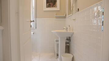 Un baño estrecho con azulejos blancos, un lavamanos pedestal y un pequeño cubo de basura.