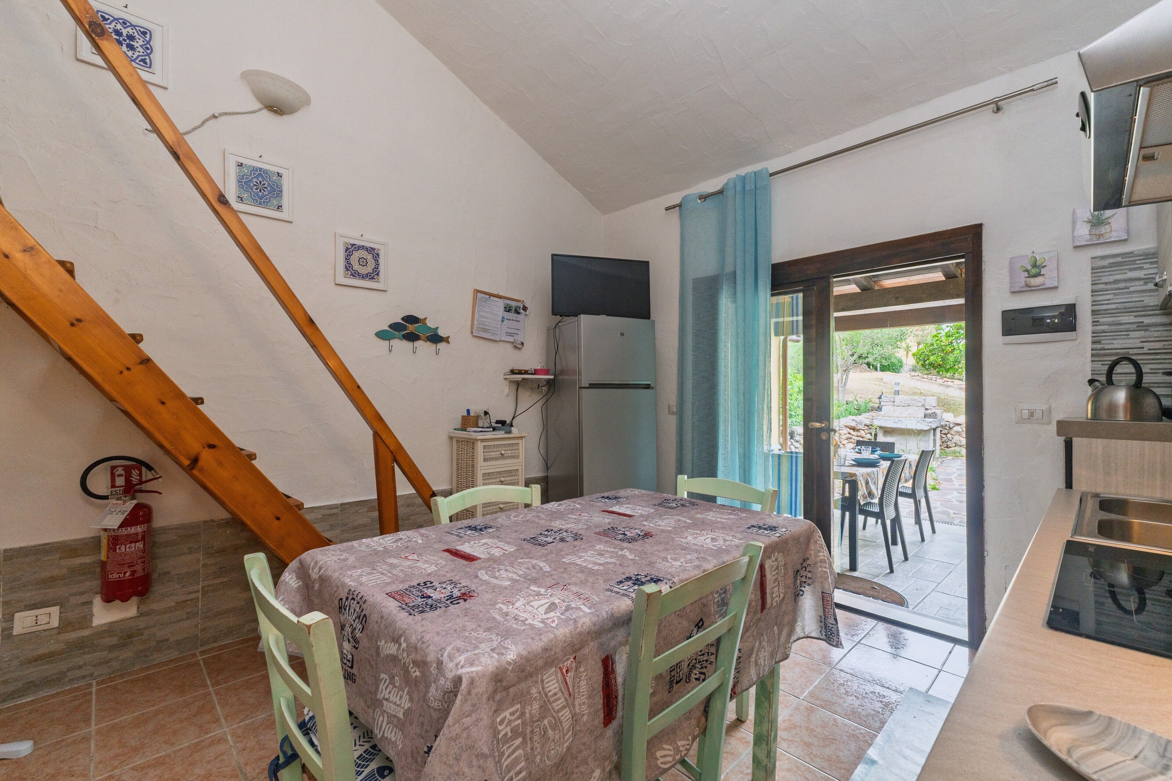 Un coin repas avec une table recouverte d’une nappe à motifs, des chaises vertes, un escalier en bois, un extincteur et un téléviseur fixé au mur.