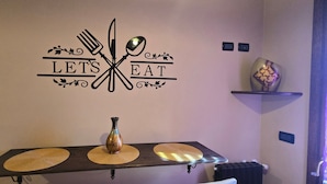 Una pared con un letrero que dice 'LET'S EAT', una mesa con individuales, un florero y un adorno en una estantería.