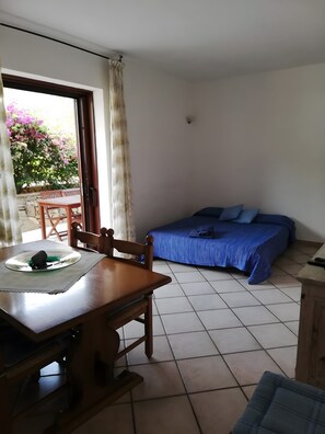 Ein Zimmer mit einem Bett, einem Tisch mit einem Teller, Stühlen, einem Fenster mit Vorhängen und Blick auf Grünfläche.