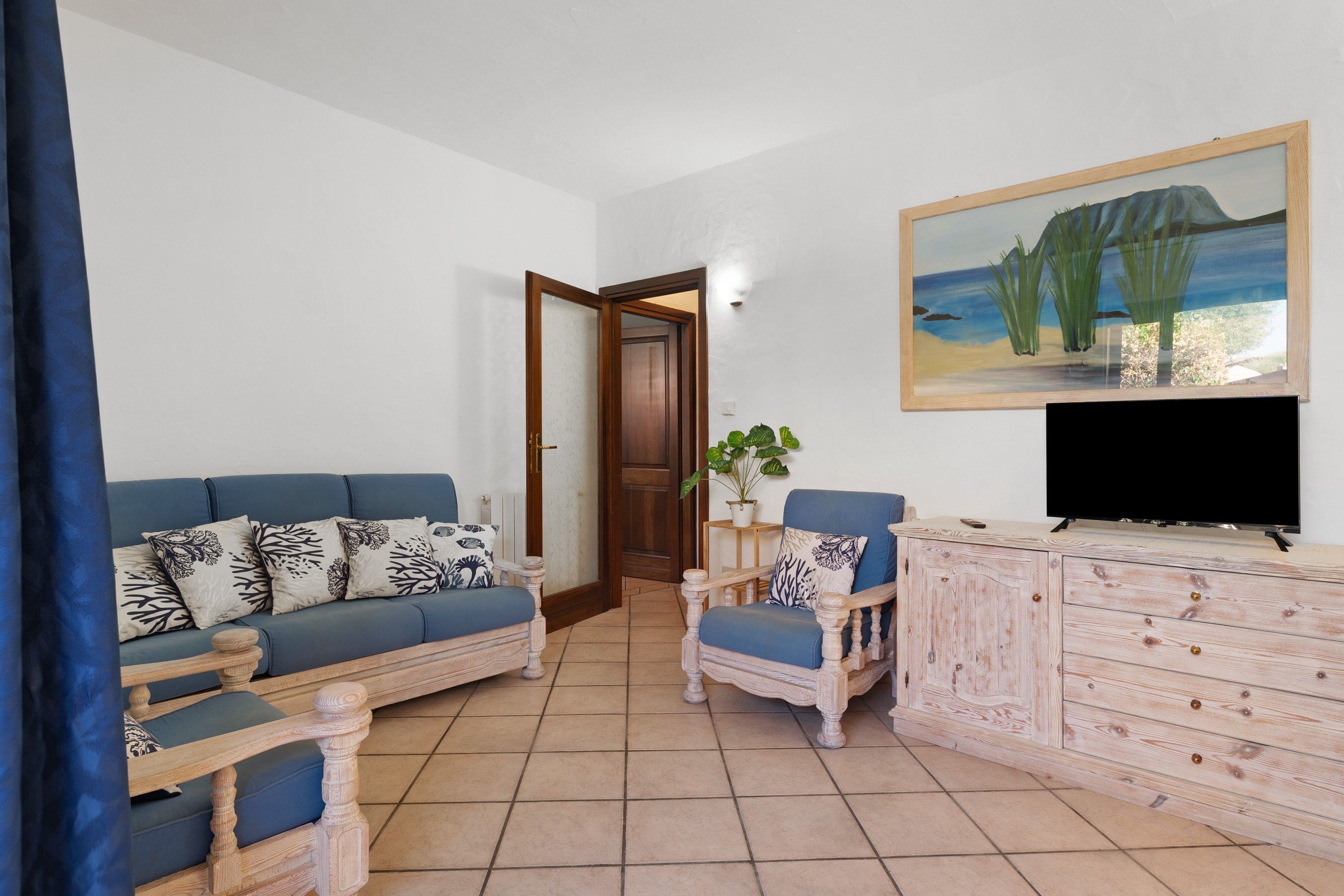 Ein Wohnzimmer mit einem blauen Sofa, einem Holzstuhl, einem Fernseher und einem Gemälde einer Strandansicht.