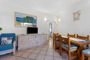 A living room with a blue sofa, a wooden dining table with chairs, a television on a cabinet, and a painting on the wall. - Holiday Home 'Villa Oliva Pittulongu' with Sea View, Wi-Fi and Air Conditioning (Pittulongu)