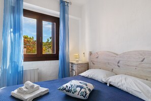 Un dormitorio con cama, ventana con cortinas, mesita de noche con lámpara, y vista a árboles desde el exterior.
