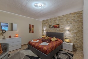Une chambre avec un mur en pierre, un lit recouvert d’une couvre-lit à motifs, des lampes de chevet, une table de chevet, une commode et un miroir.