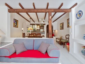 Ein Wohnzimmer mit grauem Sofa, roten Kissen, Holzbalken, einem Essbereich mit Stühlen und Tisch sowie einer Küche im Hintergrund.