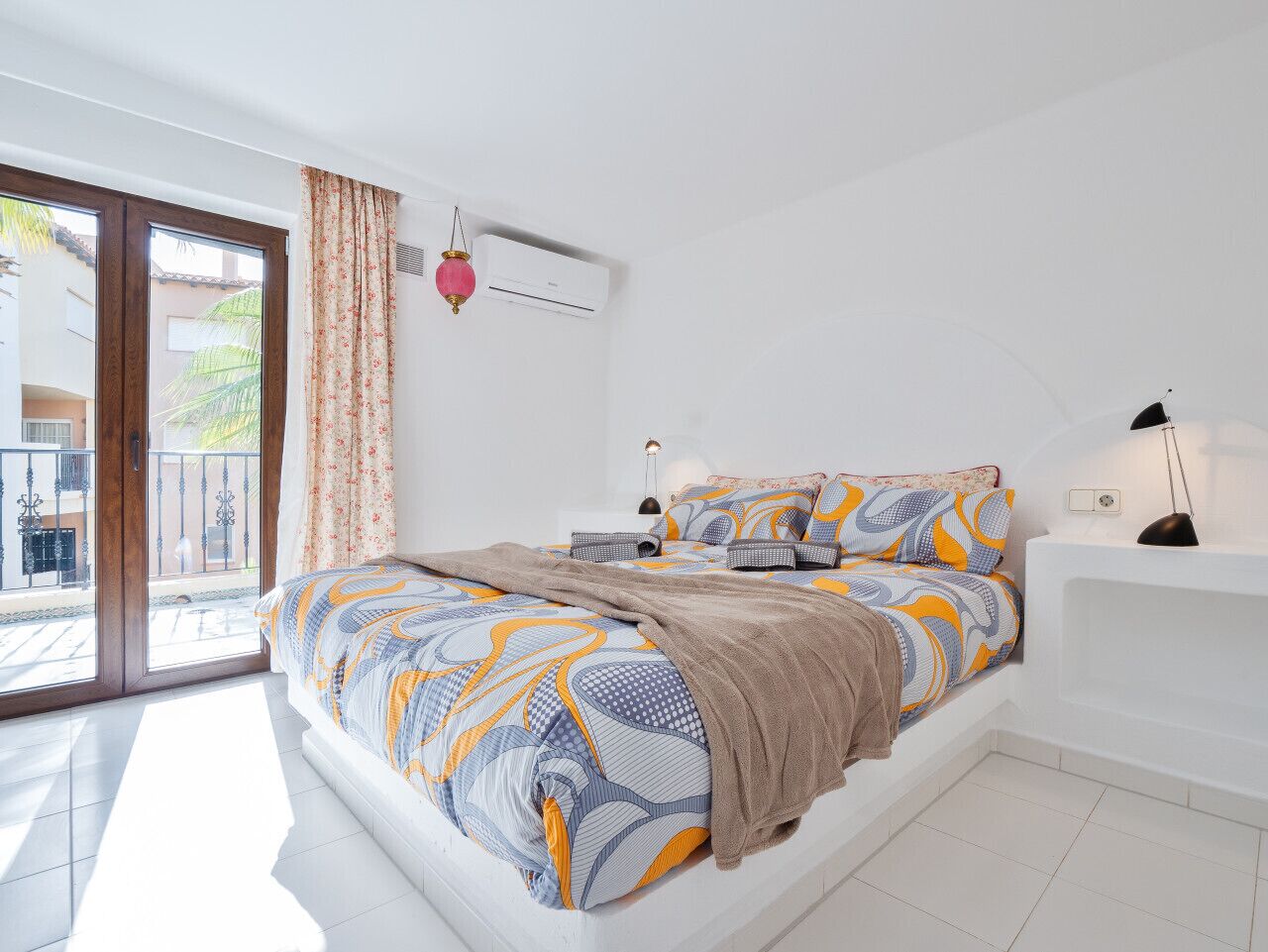 A bedroom with a large bed, a white nightstand, a white sofa, a window with curtains, and an air conditioning unit.