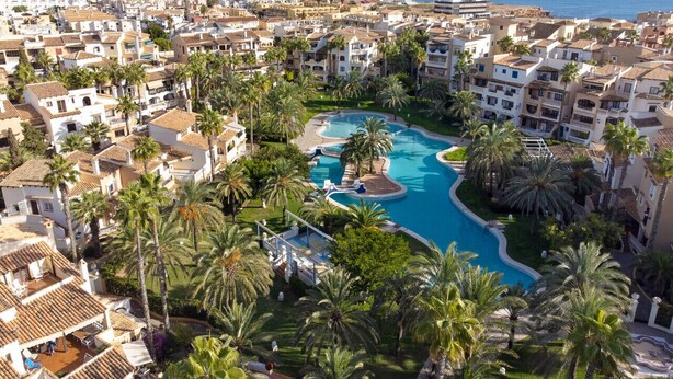 A resort with a large swimming pool surrounded by palm trees and Mediterranean-style buildings. - Sea&Sun Premium Apartamento (Torrevieja)